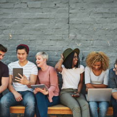 An image of six people of varying ethnicities and genders sitting on a bench seat in front of a grey brick wall looking at laptops and mobile devices.