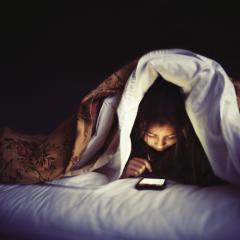 Image of a child using a mobile device, hidden under blankets on a bed in the dark.